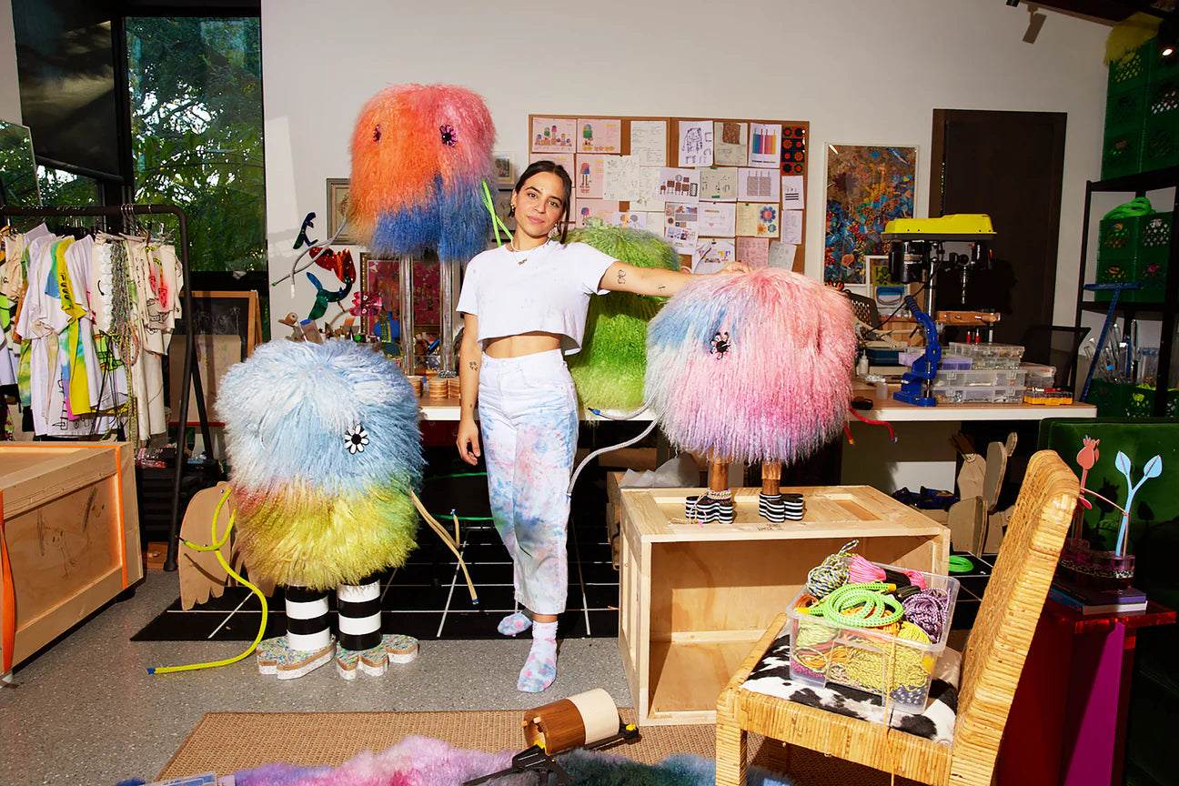 photo of artist Gabriela Noelle with her sculptures from the "imaginary friends" series in her studio. On her left, she's touching the top of the smallest imaginary friend that has pink and blue fur, on her right another imaginary friend with blue, green and pink fur and black and white stripped legs, behind her on her right an imaginary friend with pink, orange and blue fur over a table and clear acrylic legs, behind her on the left, the body of an imaginary friend over a table in green.